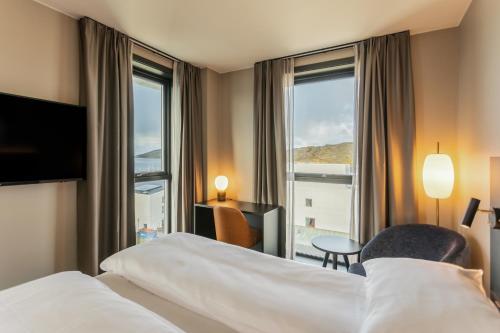 Hotel room with two white beds, a desk by large windows, and a seating area overlooking a water view and hills outside.
