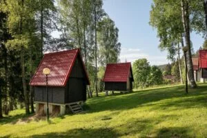 Three small black cabins with red roofs on a grassy hillside among tall trees, with a lamp post in the foreground.