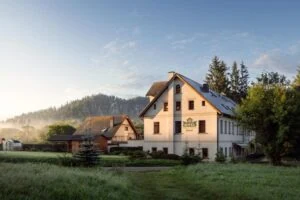 White multi-story hotel with a steep roof in a quiet countryside at sunrise, surrounded by green fields and trees.