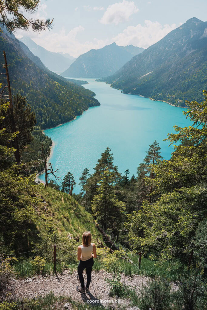 A woman stands on a trail overlooking a stunning turquoise lake surrounded by lush green forests and towering mountains under a partly cloudy sky.