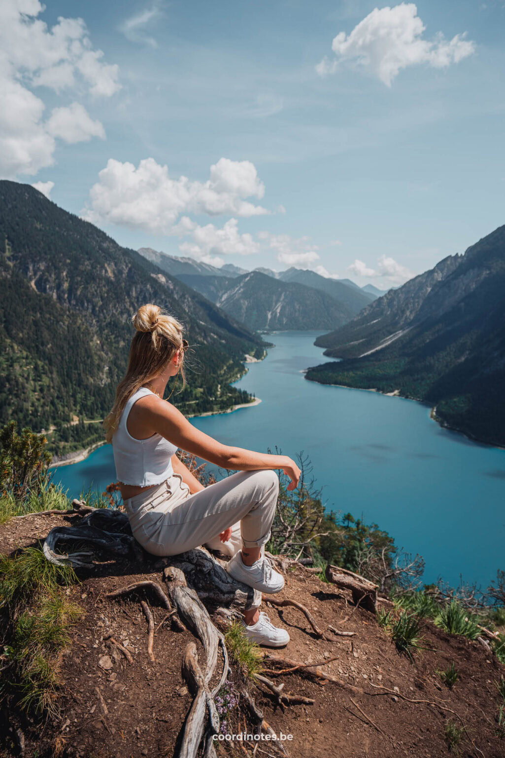 Sarah sitting on tree roots on top of a mountain watching over Plansee surrounded by mountains in the background.