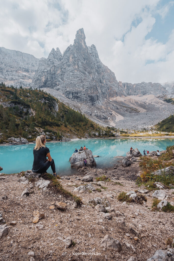 Sarah sitting on a rock in front of the blue water of Lago di Sorapis with dramatic mountain peaks in the background