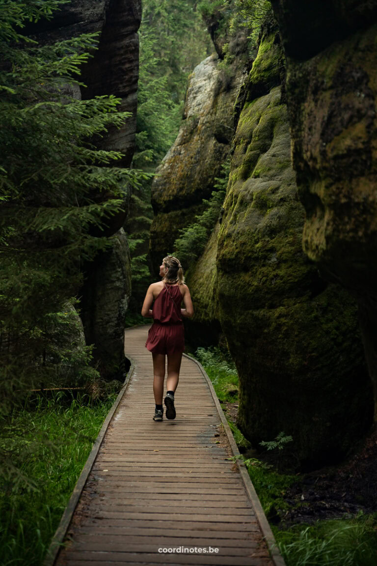 Woman in a burgundy romper walking along a wooden boardwalk between mossy rock walls in a forest canyon.