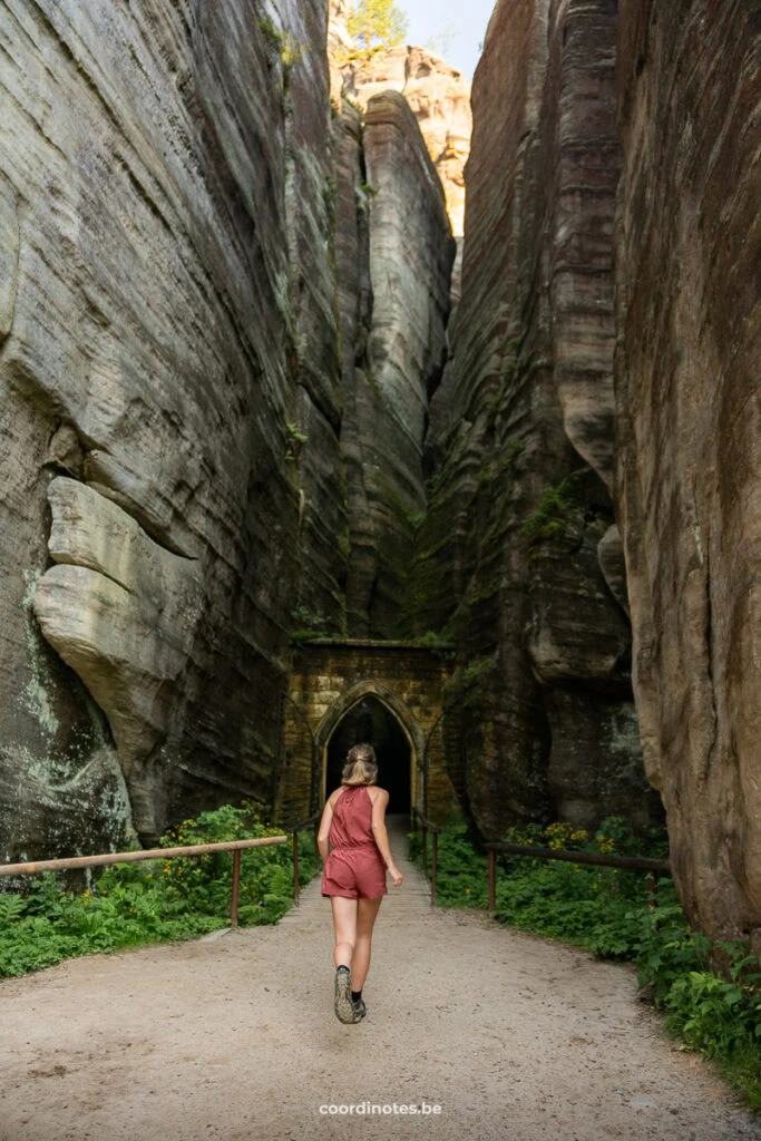 Woman in a pink romper walks toward a stone archway between towering canyon walls.