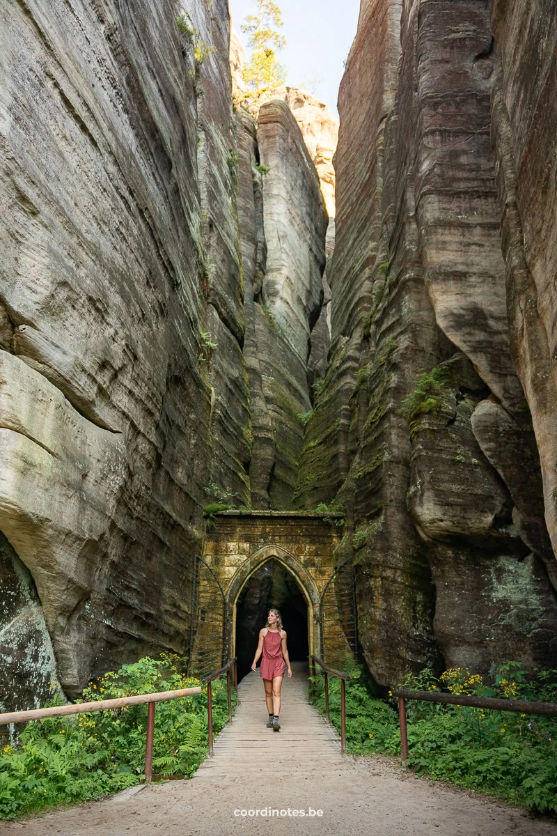 Woman in a pink romper walks along a wooden bridge toward a stone arch doorway between towering canyon walls.