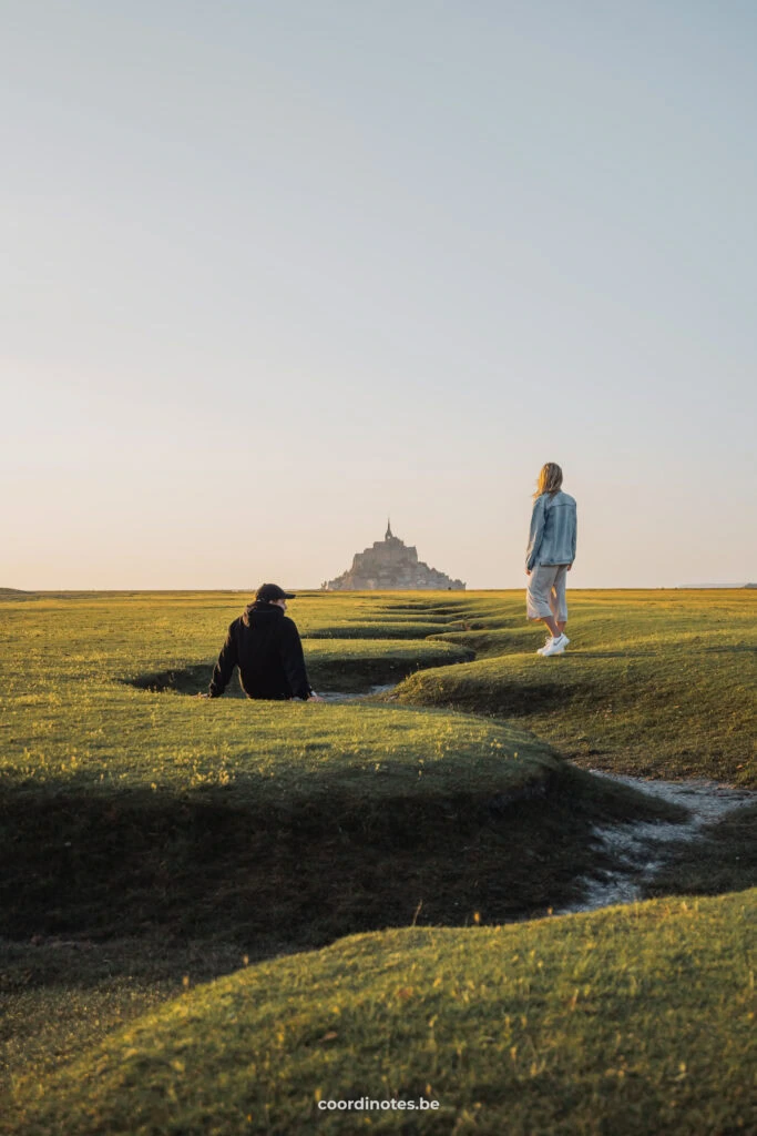 A dried up meandering stream towards Mont Saint-Michel in the background. This is a castle on a small island. Cédric is sitting on the edge of the stream and Sarah is standing next to it.