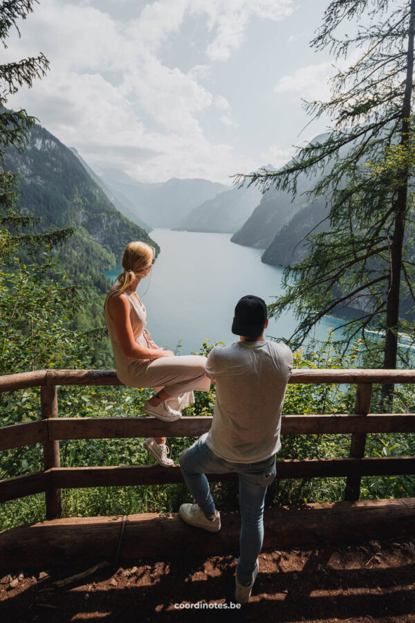 Sarah sitting on the fence and Cédric standing against it while watching over Königssee in the distance, surrounded by mountains from the viewpoint of Rabenwand