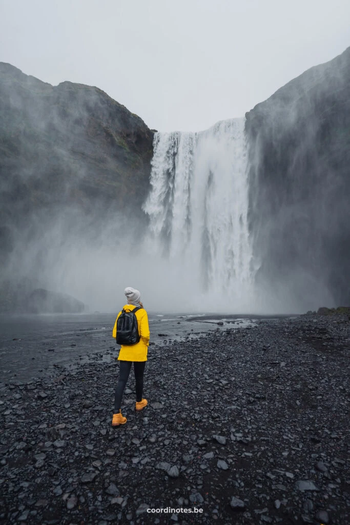 Sarah walking towards the huge Skogafoss, a big waterfall tumbling down a big black wall. Sarah is wearing a bright yellow jacket.