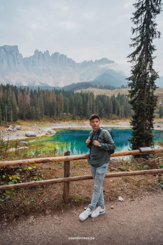 Cédric aan het hek aan Lago di Carezza​ met naaldbomen en bergtoppen in de achtergrond