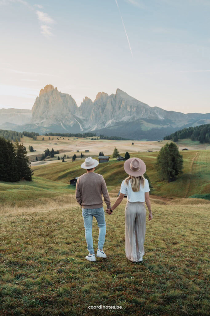 Wij twee die hand in hand wegwandelen op het gras met de grote bergweide van Seiser Alm en een grote bergketen op de achtergrond tijdens zonsopgang.