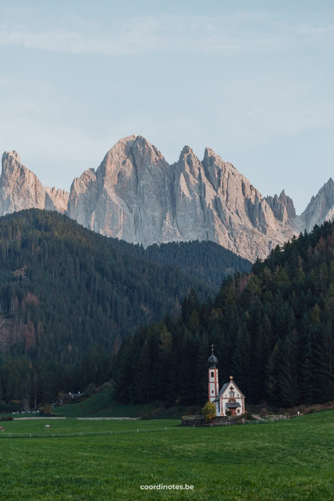 De kerk van St. Magdalena in een weiland met gras voor een bos met pine trees en dramatische bertoppen op de achtergrond in Val di Funes