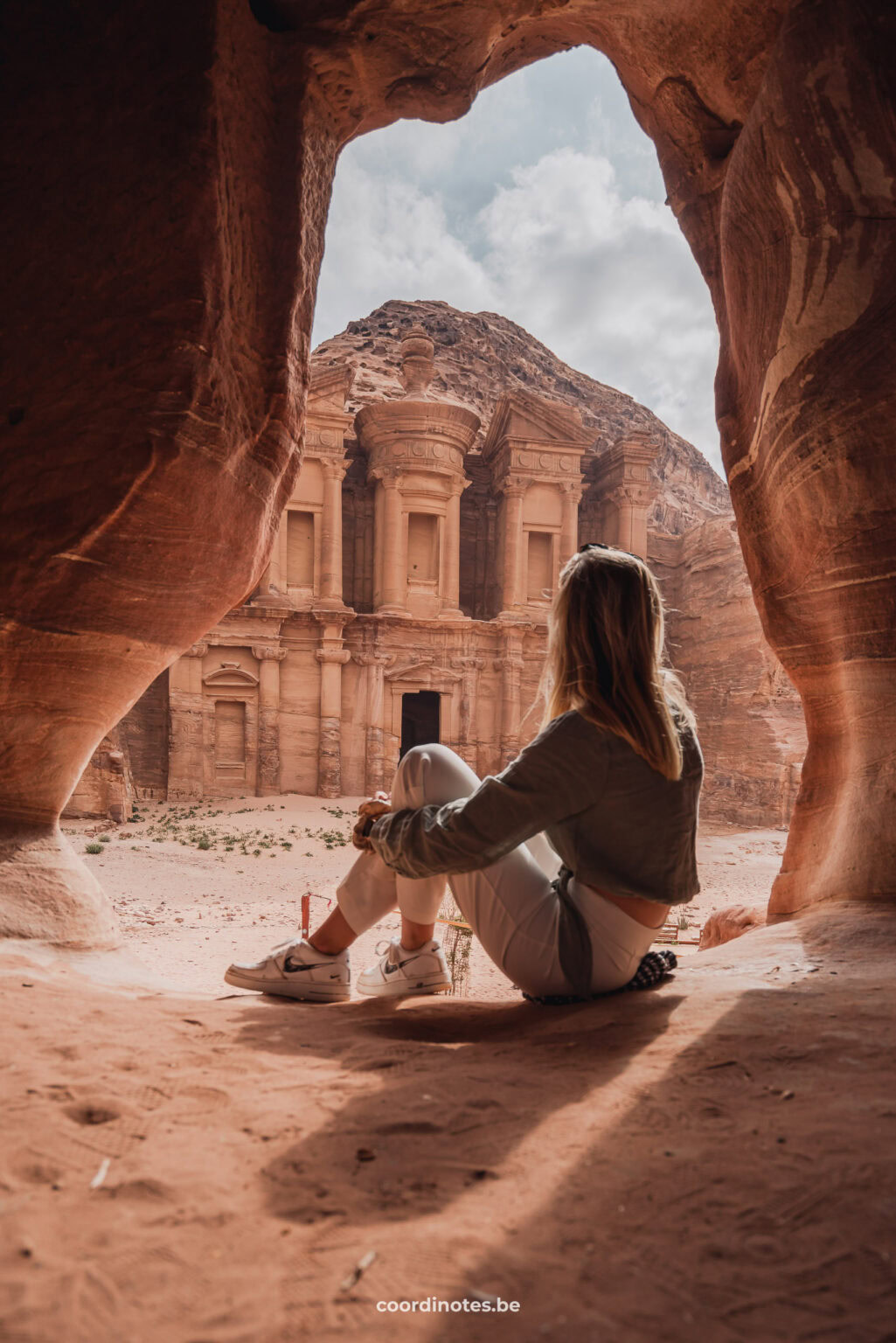 Sarah sitting in a cave, watching over The Monastery, a big building with columns carved in the orange rocks in Petra.