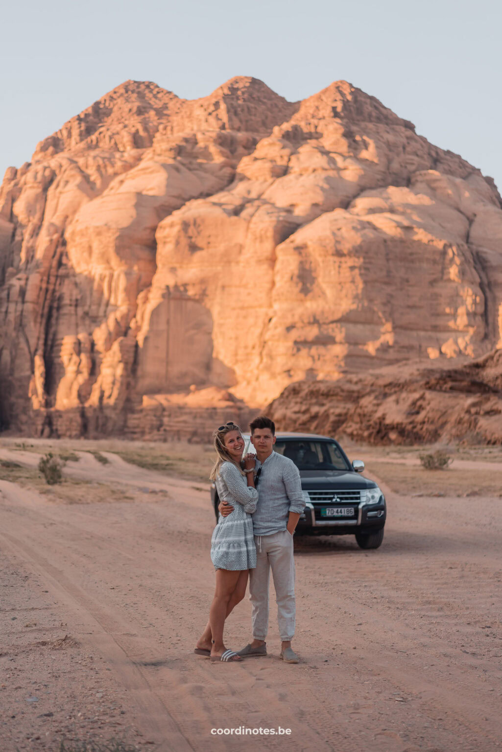 Us in the Wadi Rum desert in front of our car with a huge rock formation in the background during sunset