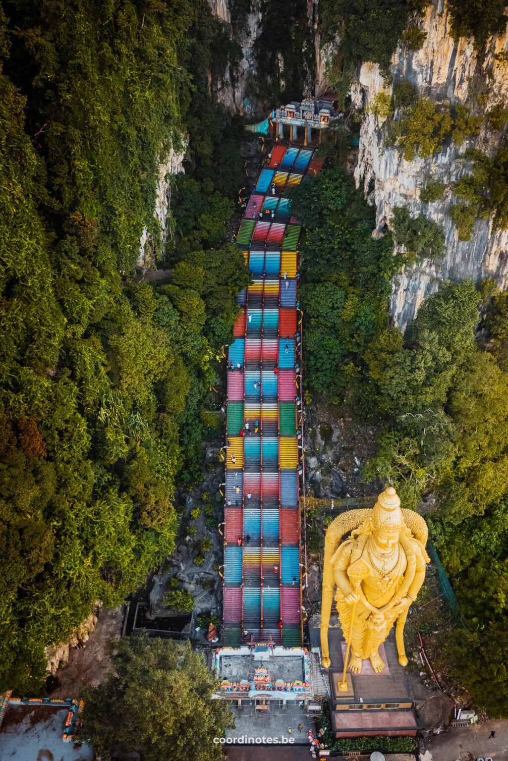 Top view of a long colorful stairs surrounded by trees leading to the Batu Caves in the limestone mountains, and a huge golden statue of Lord Murugan, the Hindu god of war at the bottom of the stairs.