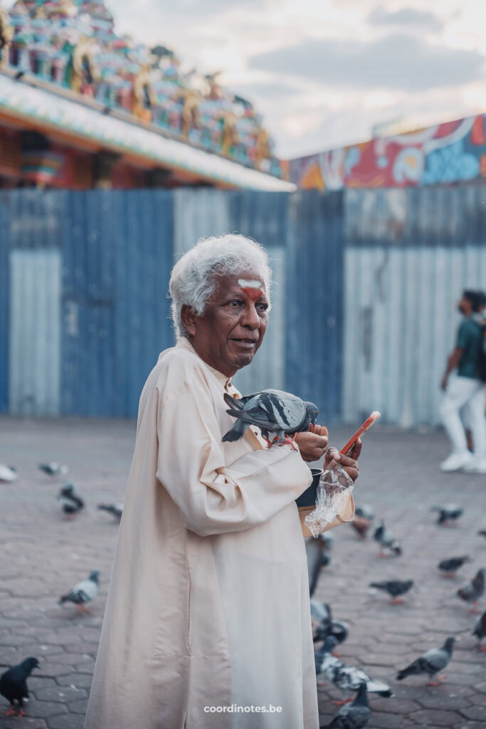 A hindu man holding a pigeon, a bag with seeds and his phone, surrounded by more pigeons on a square.