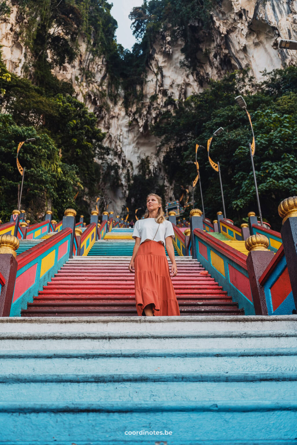 Bottomview of Sarah walking down a rainbow stairs at Batu Caves with huge limestone rocks at the top of the stairs