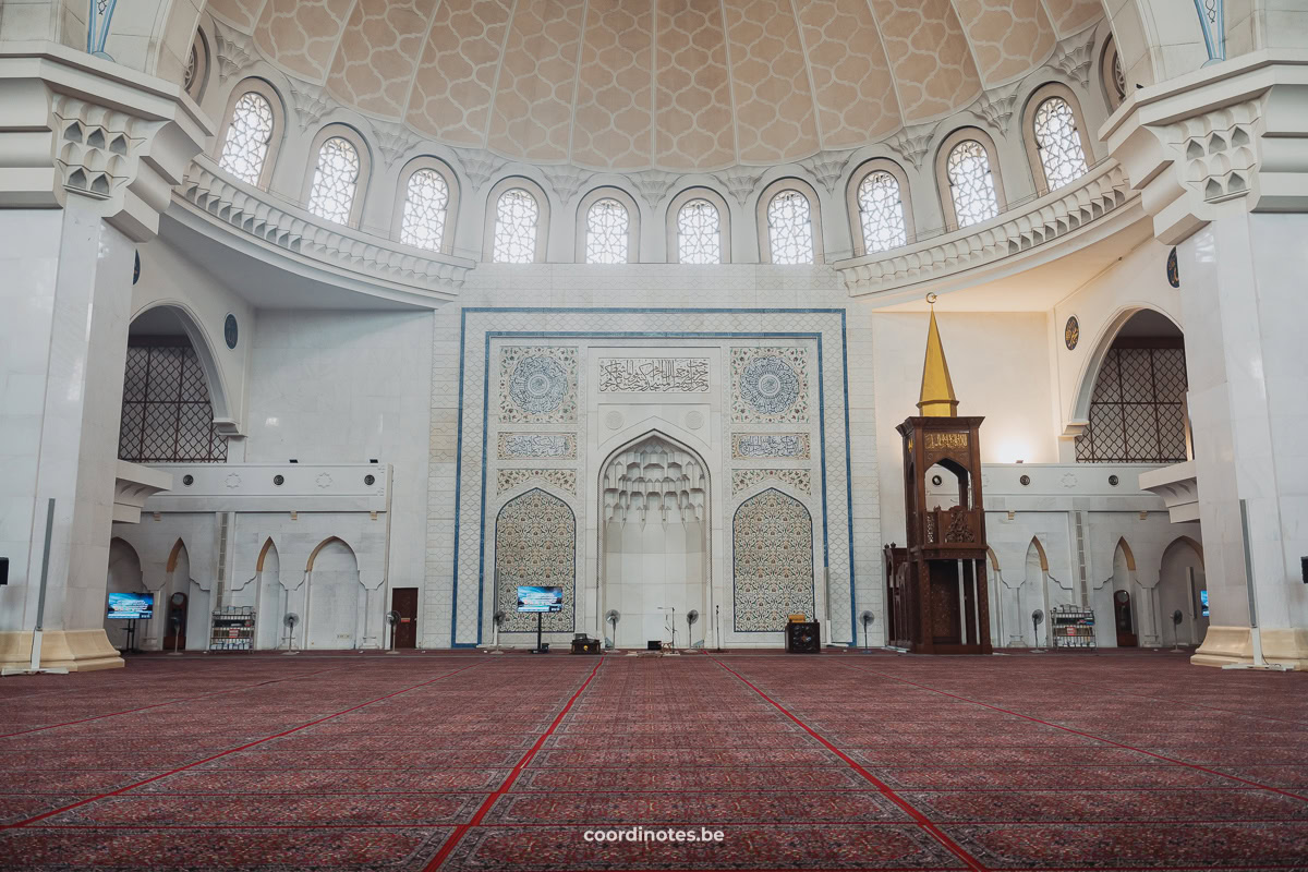 Inside the prayer hall of the Wilayah Persekutuan Mosque with red carpets on the floor and a niche in the wall under the large dome.