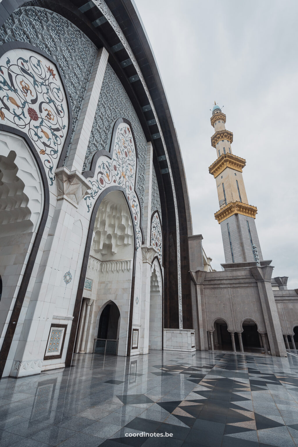 A wall of the Wilayah Persekutuan Mosque with arches, decorated with white and blue mosaic and a golden minaret in the background on the right.