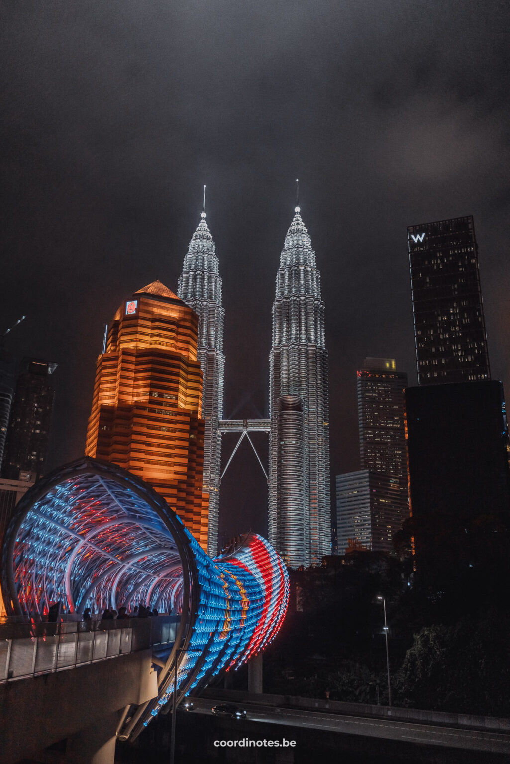 The illuminated Saloma Link bridge in the colors of the Malaysian flag with another yellow illuminated sky scraper behind and the two white illuminated Petronas Twin Towers in the background amidst a few other skyscrapers at night in Kuala Lumpur.