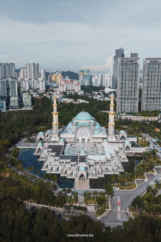 A big white mosque with blue domes and two big minarets surrounded by trees and bushes with the city of Kuala Lumpur in the background.