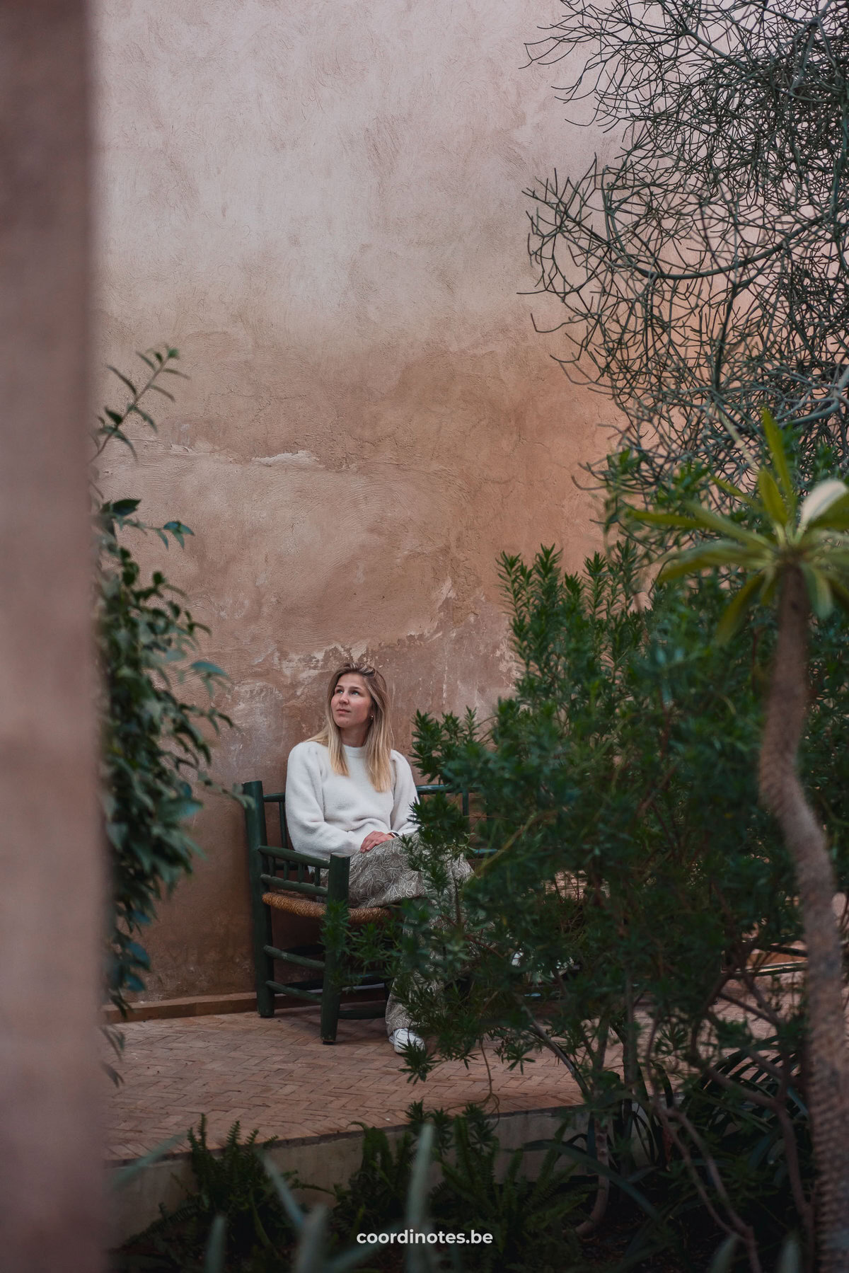 Sarah die op een bankje zit in Le Jardin Secret voor een oranje muur tussen groene planten