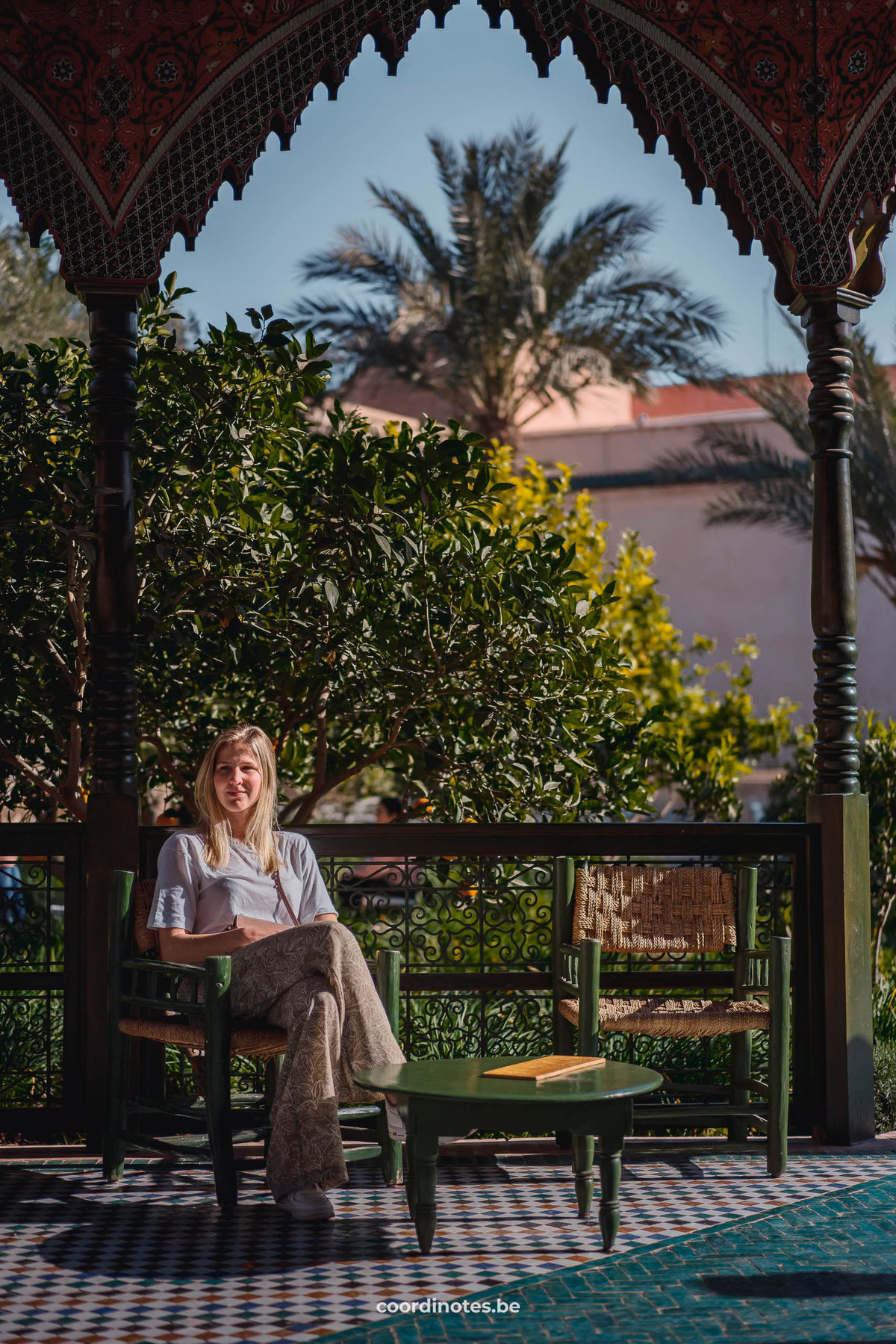 Sarah die op een stoel zit naast een tafeltje en een andere stoel in Le jardin Secret onder het paviljoen met planten en een palmboom in de achtergrond.