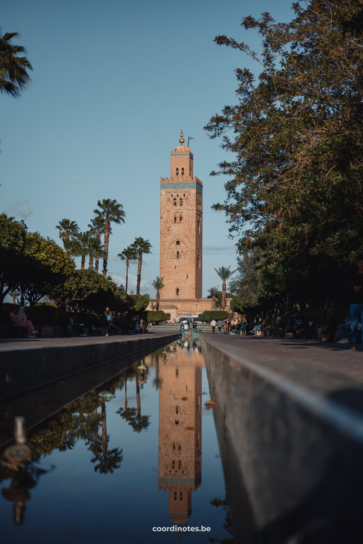 De grote oranje toren van de Koutoubia Mosque en de weerspiegeling ervan in een grote vijver ervoor met ernaast bomen en palmbomen.