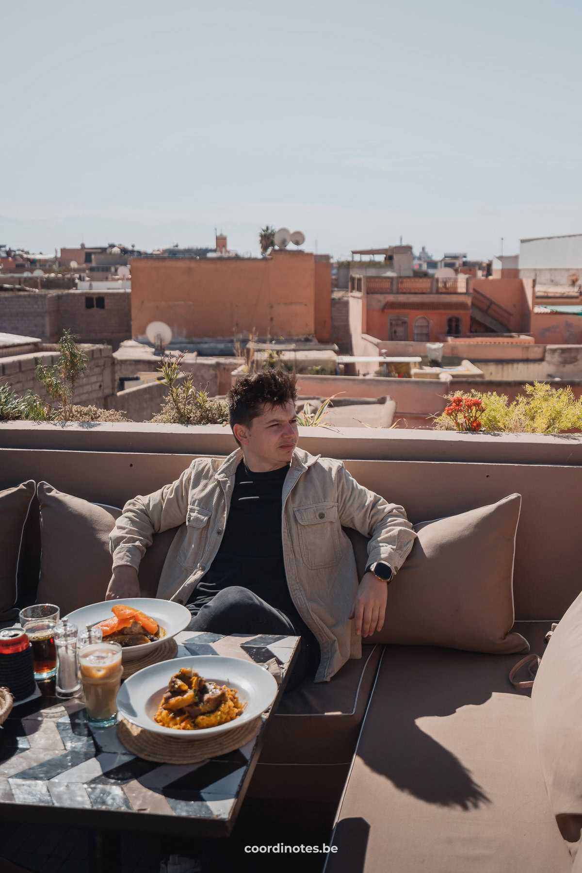 Cédric in een lounge in een rooftop bar met uitzicht over andere daken in Marrakech met twee borden met eten op tafel voor hem.