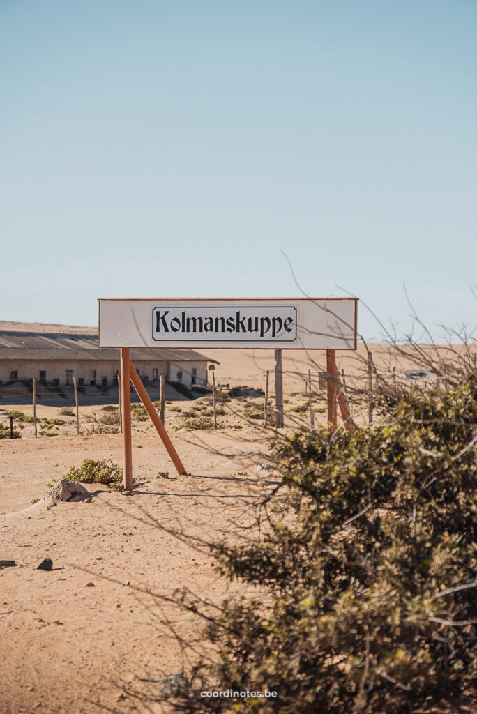 A wooden sign with 'Kolmanskuppe' in the sand with some bushes around.