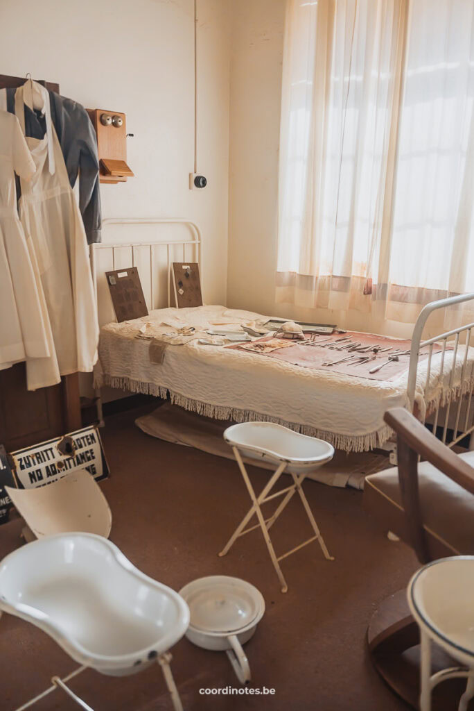 Een kamer in het museum van Kolmanskop met een ziekenhuis bed, enkele doktersjassen die aan de muur hangen en verschillende baby badjes. Op het bed liggen verschillende ziekenhuis materialen.