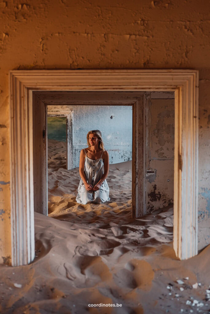 Sarah die op haar knieën zit in het zand in de deuropening in één van de verlaten huizen in Kolmanskop die voor de helft gevuld zijn met zand.