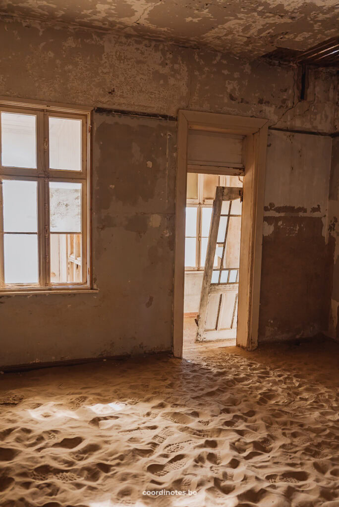 An abandoned room in Kolmanskop with a broken door and the floor covered in sand.