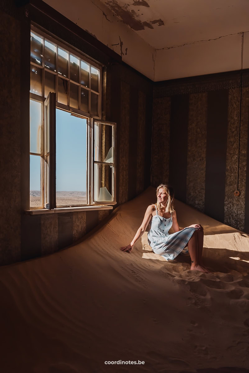 Sarah die op een berg zand zit in een verlaten huis in Kolmanskop. Ze zit naast een gebroken raam waar de zon door naar binnen schijnt.