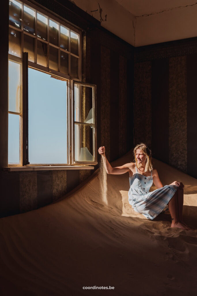 Sarah sitting on a pile of sand, letting sand slippin through her fingers next to a broken window inside an abandoned house in the ghost town of Kolmanskop.