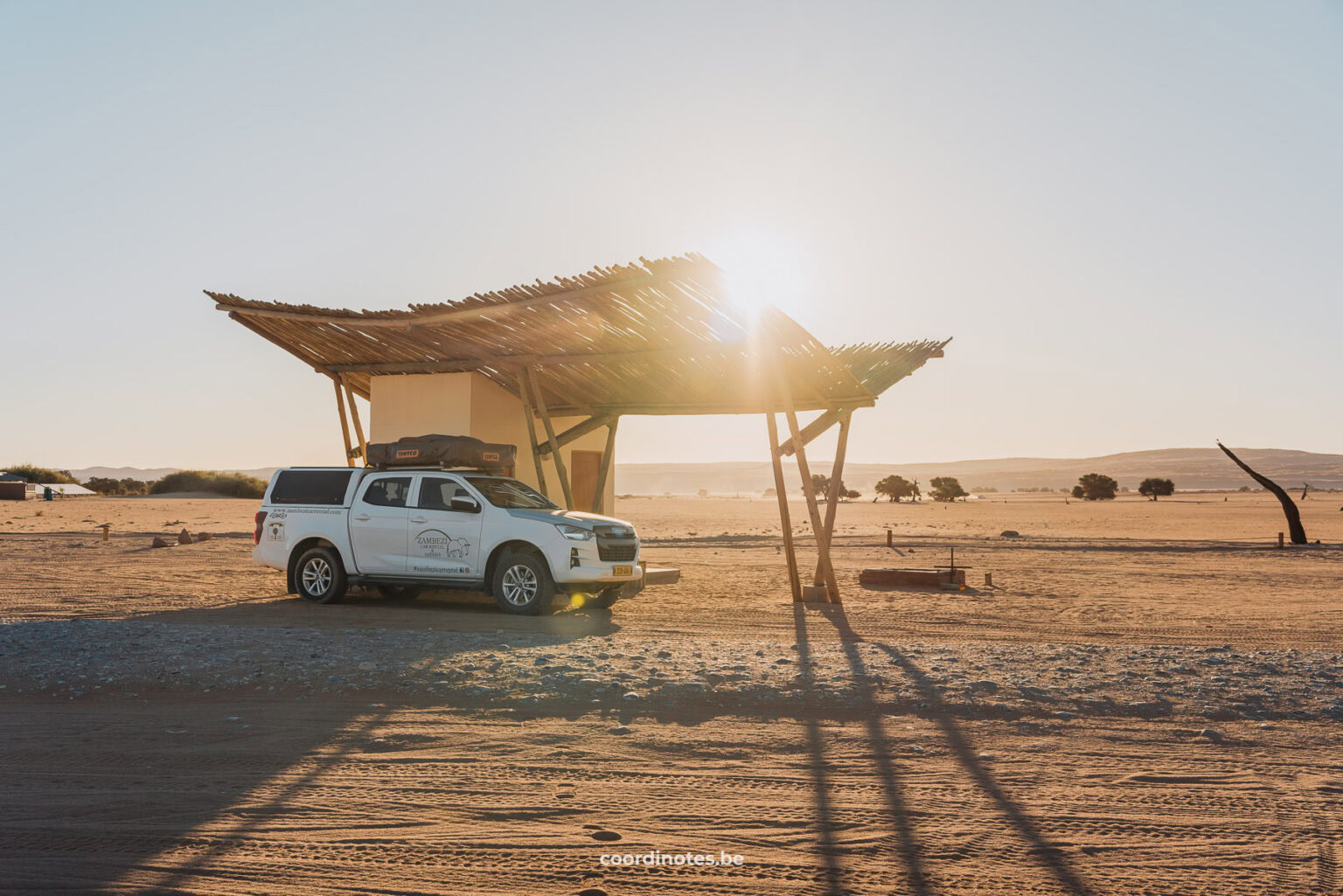 Our pick-up truck standing next to a little building with canopy. In the background you see the desert of Sossusvlei and the sun is shining right over the canopy at sunset.