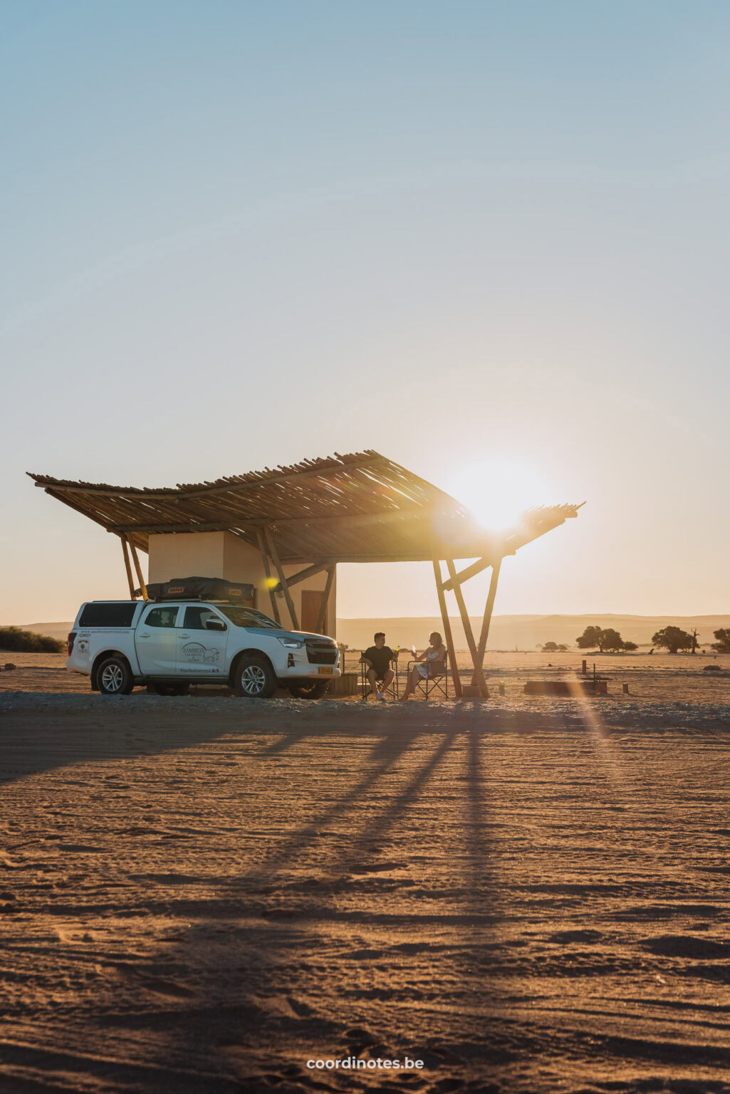 The two of us sitting under the canopy, next to a little building , of our camping pitch in Sesriem Oshana Campsite and our white pick up truck with the sunset in the background