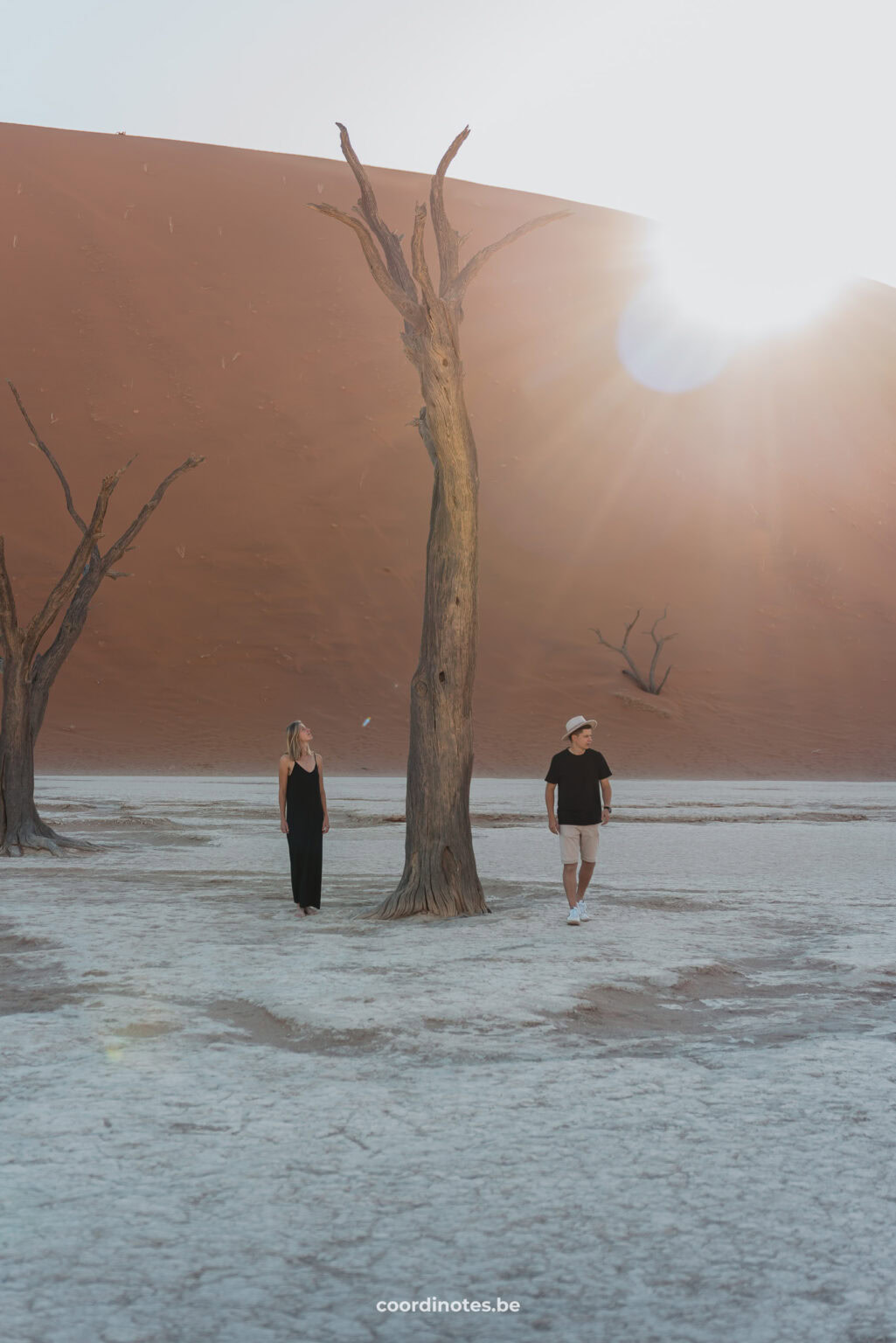 The two of us standing on both sides of a dead tree on a white clay ground. In the background you see a red sand dune and the sun shining right over the sand dune.