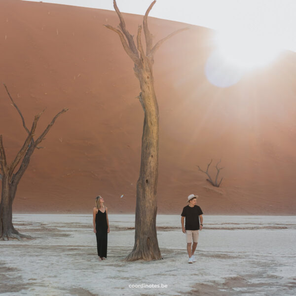 Wij twee met een dode boom die tussen ons staat op de witte kleigrond van Deadvlei. Op de achtergrond zie je een rode zand duin en de zon die er net over schijnt waardoor er een flair in de lens ontstaat.