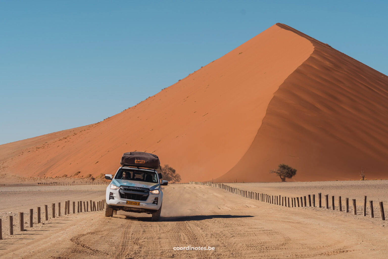 Our white pick-up truck driving away from a big sand orange sand dune over a sand road towards the camera.