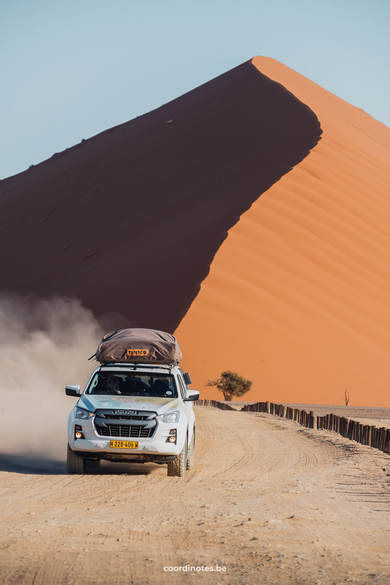 Our white pick-up truck driving in front of a big sand dune. The right side of the sand dune is illuminated by the sun, while the left side is in shade.