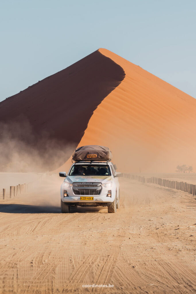 Our white pick-up truck driving in front of a big sand dune. The right side of the sand dune is illuminated by the sun, while the left side is in shade.