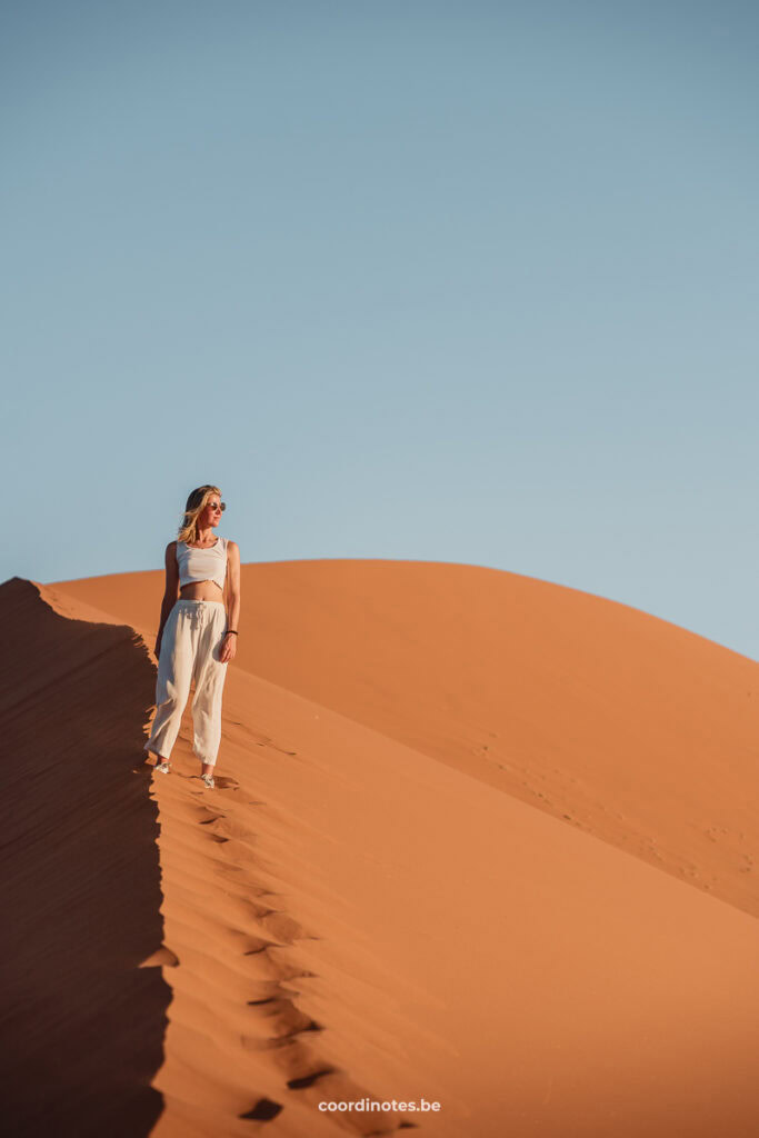 Sarah standing on Dune 45, a sand dune that is illuminated at the right side by the sun and has shadow on the left side. In the background you see the top of the dune and in the foreground you see Sarah's footsteps towards where she is standing.