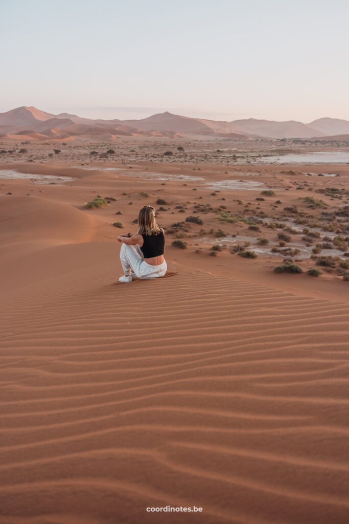 Sarah sitting on an orange sand dune watching over a plane with more big sand dunes in the background.