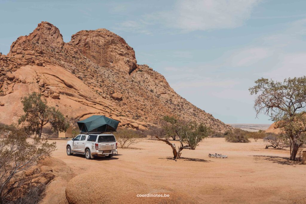 Our white pick-up truck with open rooftop tent on a sand plane between a few bushes with a large granite mountain peak on a campsite of Spitzkoppe in the background.