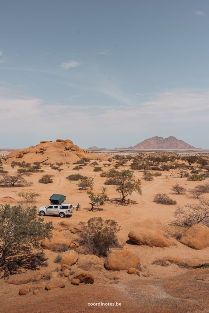 Onze witte pick-up truck met open daktent in een grote zandvlakte met groene bosjes en rotsen verspreid over de vlakte. In de verte zie je een grote granieten berg.