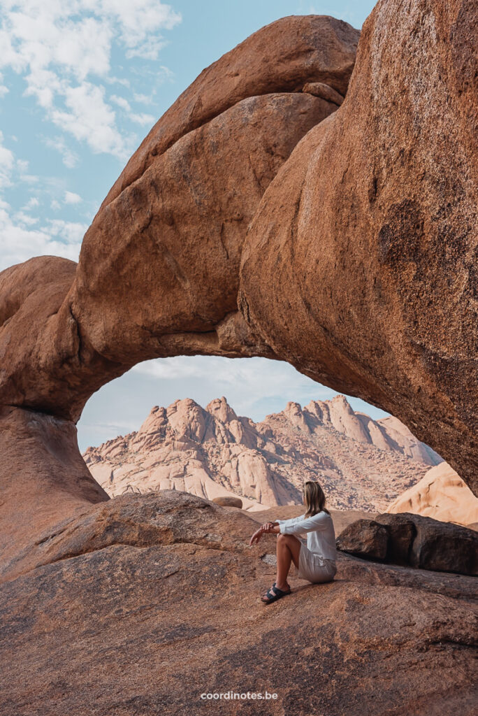 Sarah sitting on a big red rock under a natural stone bridge forming a natural frame to the red granite peaks of Spitzkoppe in the background.