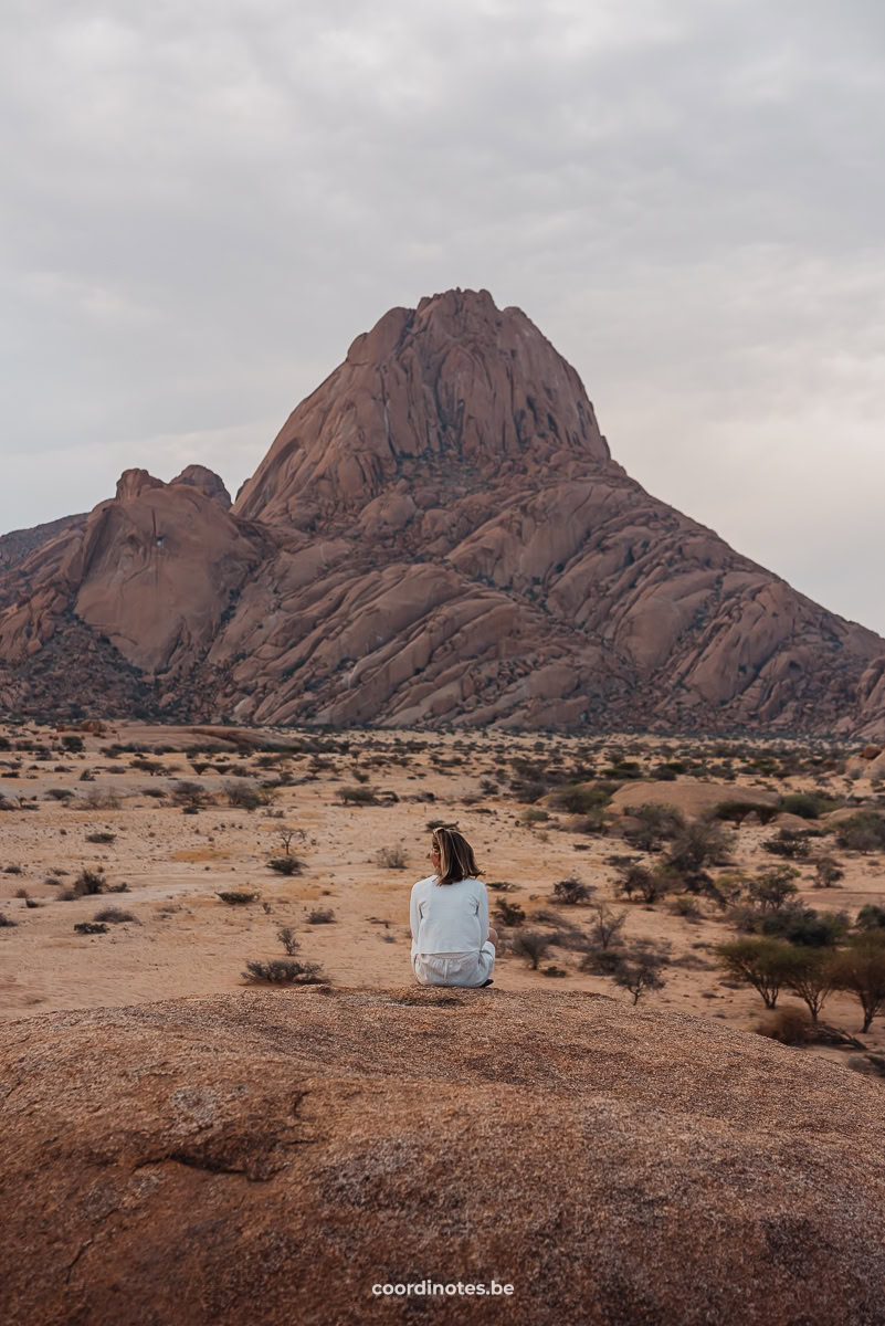 Sarah die op een rots zit met een grote zandvlakte met bosjes met daarachter de grote rode granieten bergpiek van Spitzkoppe in de achtergrond.