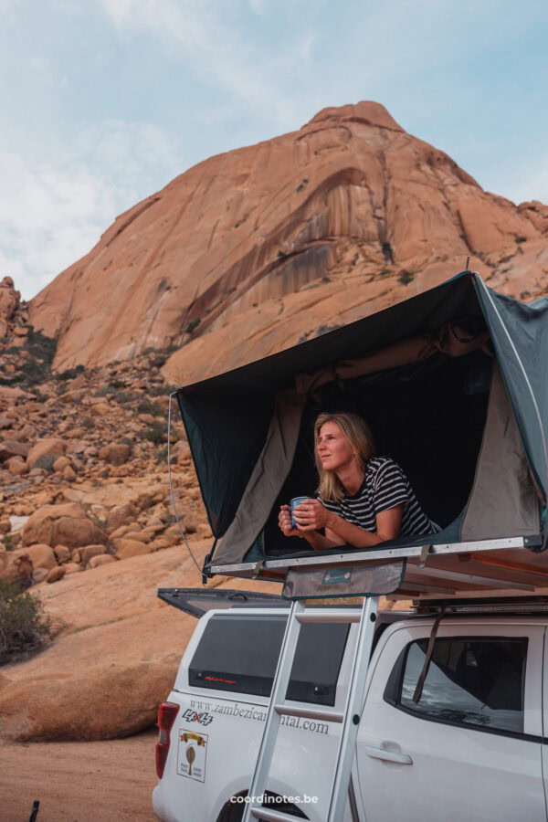 Sarah laying in the rooftent on top of our white pick-up truck looking outside holding a tea cup. In the background, you see the large granite peak of Spitzkoppe.