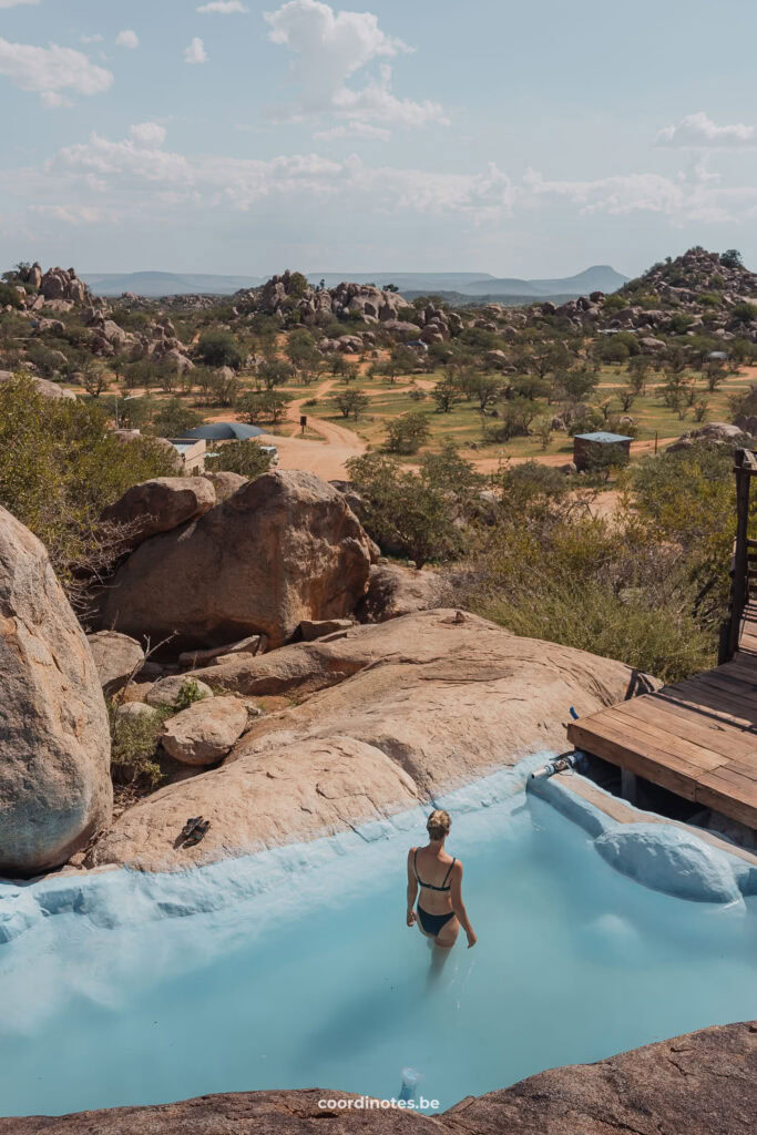 Sarah standing in a blue pool surrounded by large boulders and rock, with the view of a plane filled with trees in the distance at Hoada Campsite