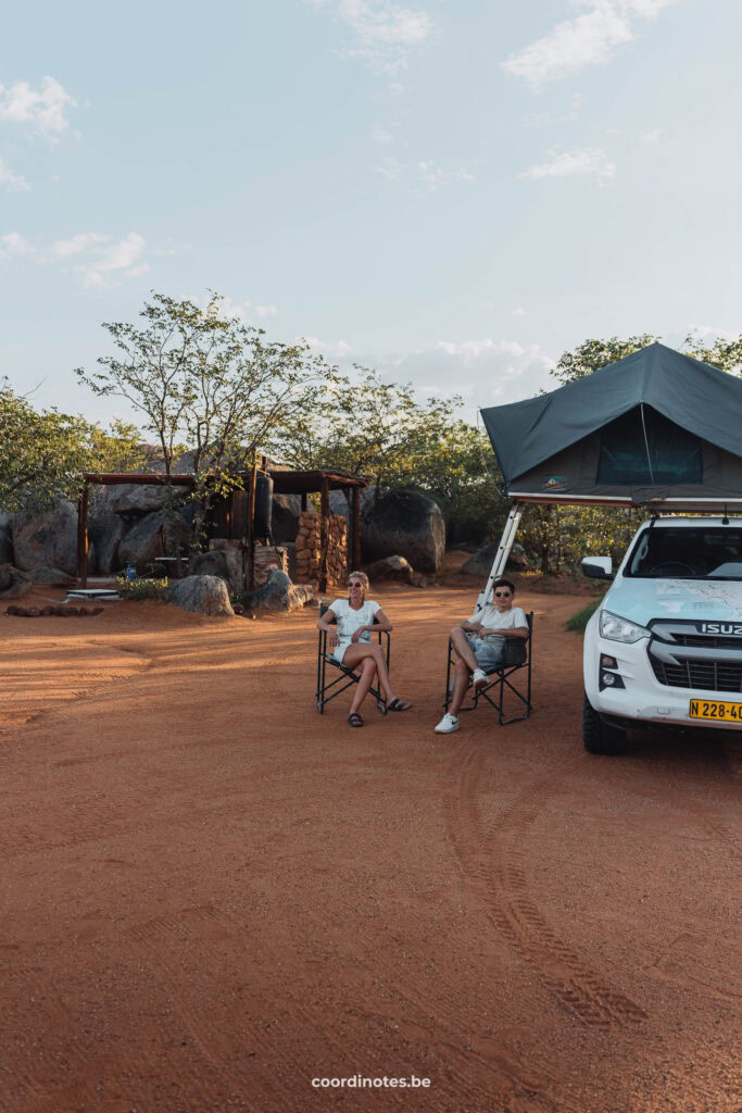 The two of us sitting in our camping chairs next to our white pick-up truck with open rooftent on red sand with our canopy and camping facilities in the background.